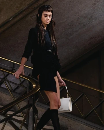 A brunette model walks down a staircase wearing a black mini dress, knee high socks, and carrying a white leather purse.