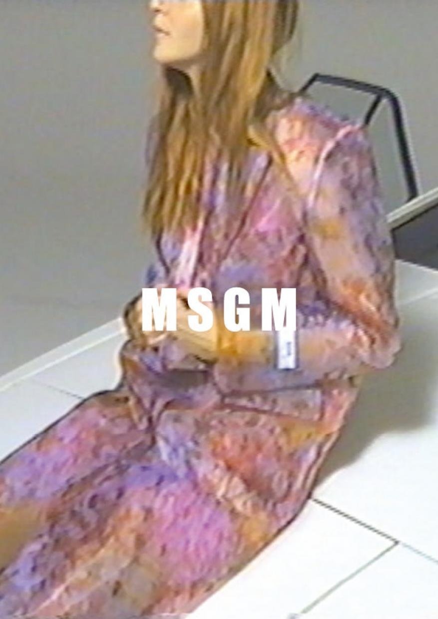 Woman in a patterned suit sitting against a gray backdrop with the logo MSGM displayed in front of her.