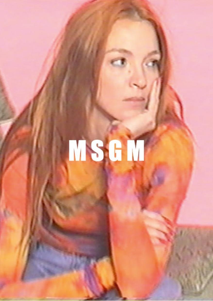 Adult woman in long-sleeve tie-dye shirt sits indoors with her head resting on her hand in a thoughtful pose against a pink background.