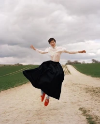 Model with short hair wearing a white ruffled blouse and black skirt jumping along a dirt path under an overcast sky