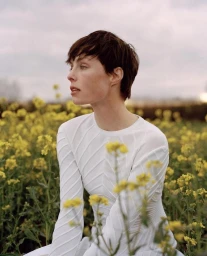 A short-haired woman in a textured white long-sleeve top sitting amongst a field of yellow wildflowers under an overcast sky.