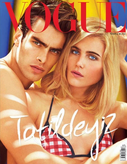 Fashion magazine cover featuring a brunette man embracing a blond woman wearing a red-and-white bikini top, with the VOGUE logo at the top.