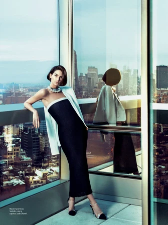 Woman in a strapless black dress and white jacket leaning against a high-rise window overlooking a city skyline