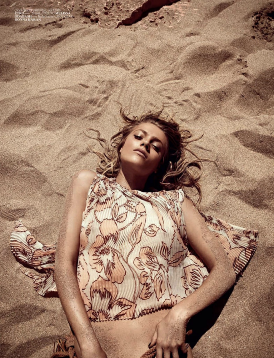 Blonde woman lies on tan sand in a floral print top, with arms at her sides, viewed from above in an eye-level full shot.