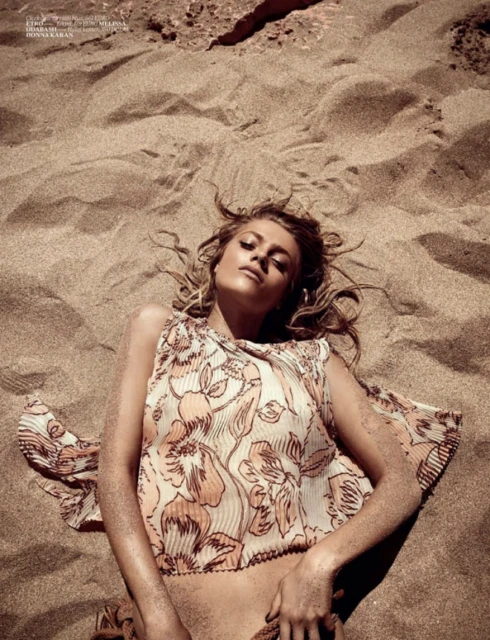 Blonde woman lies on tan sand in a floral print top, with arms at her sides, viewed from above in an eye-level full shot.