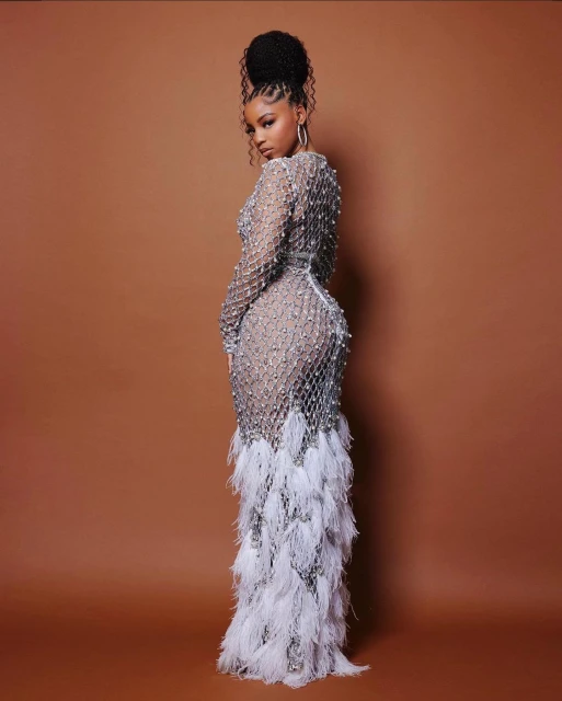 Adult female posing, looking over shoulder in a long, silver beaded gown with white feathered trim, indoor photo studio, tan backdrop.