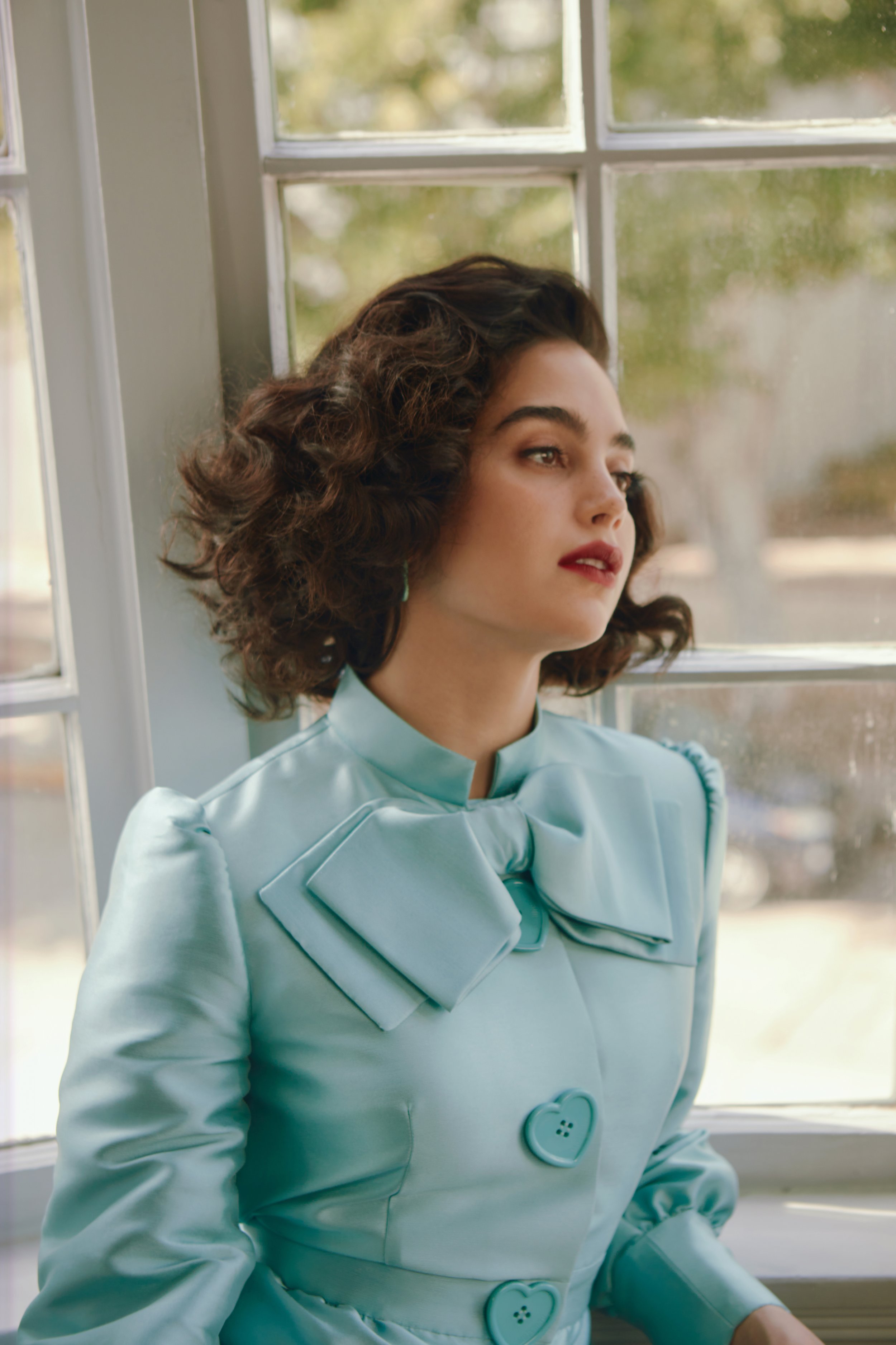 Portrait of a young woman with dark hair in a light blue jacket with heart-shaped buttons, standing by a bright window.