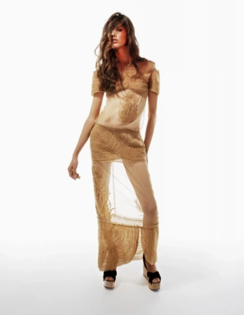 Elegant woman in sheer beige dress poses in a brightly lit photo studio showcasing flowing wavy brunette hair and stylish platform heels.