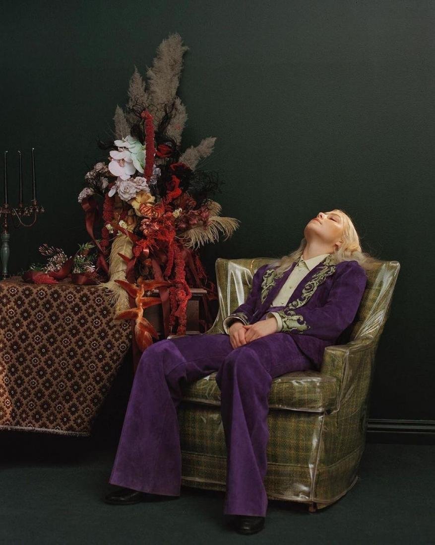 Woman with blonde hair lounges in a green upholstered chair wearing a purple embroidered suit in an indoor setting with dark green wall.