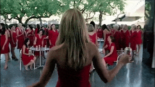 Back view of a woman with blonde hair holding a can; background of numerous women in red dresses at an outdoor event.