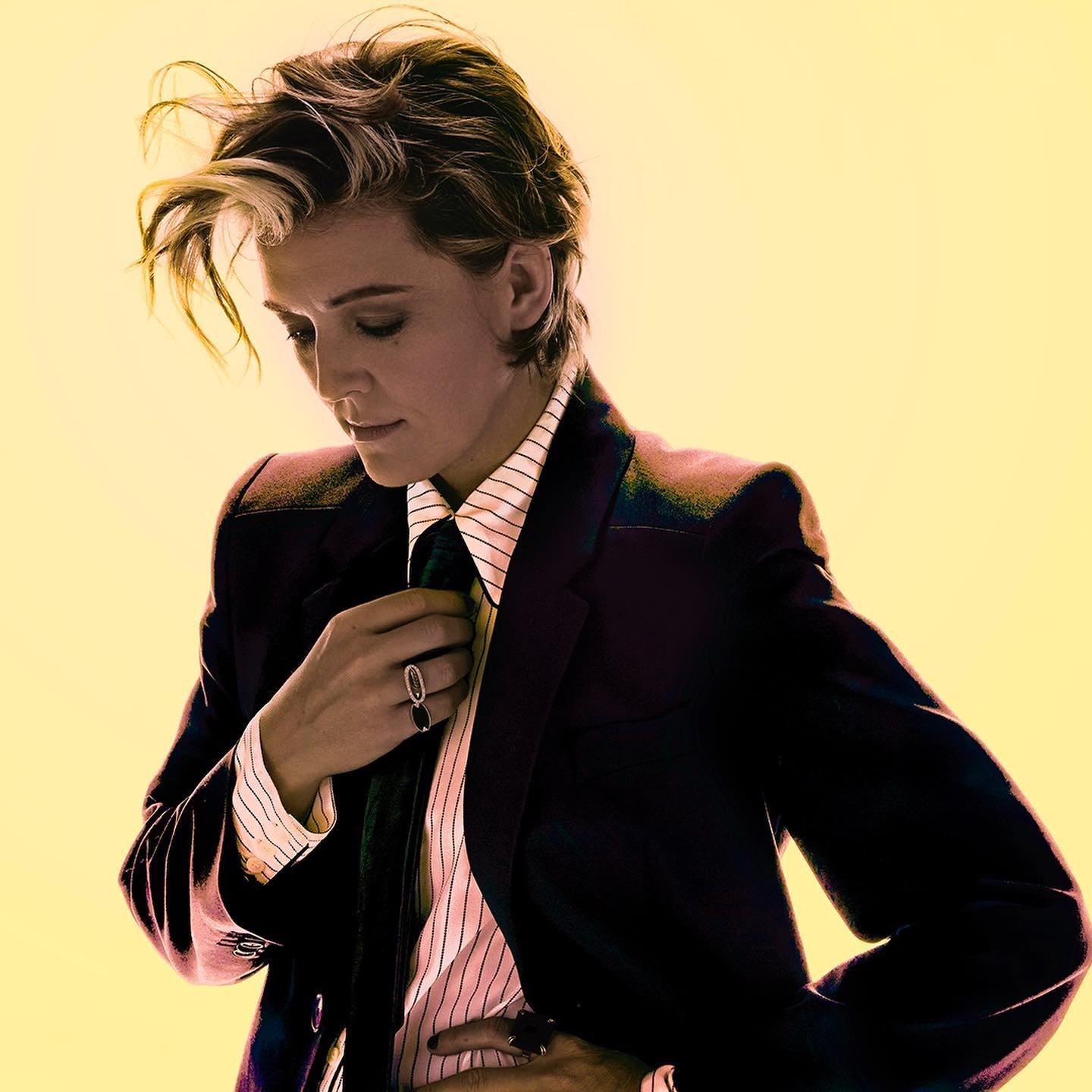 Woman in a black suit and striped shirt adjusting a dark tie, looking down with fair skin and blonde hair against a yellow background.