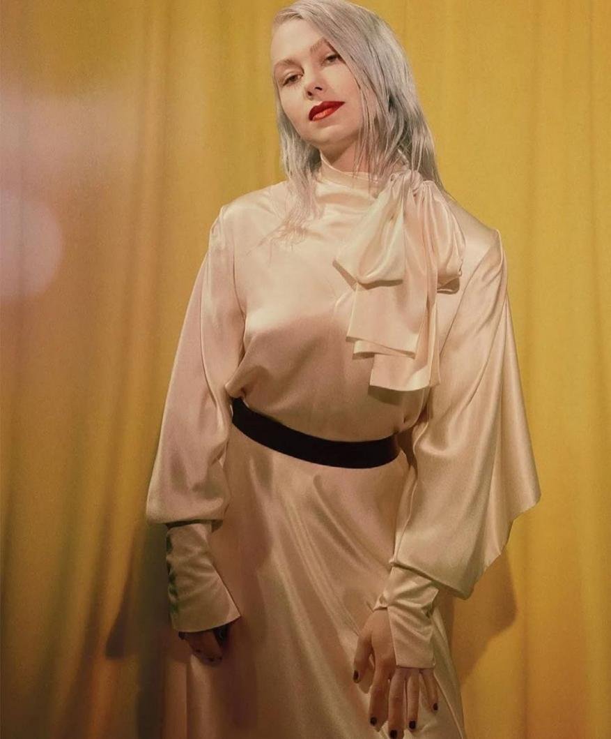 Woman with platinum hair and red lipstick wears a long, light pink dress with a black belt standing in front of a yellow curtain.
