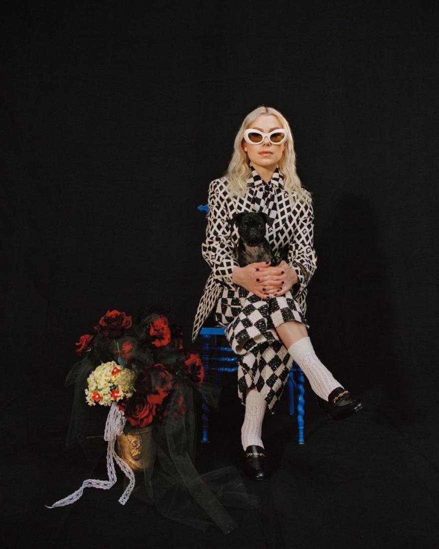 Woman in checkered suit poses with dog. Portrait in front of black background.  Bucket with roses sits on the floor.