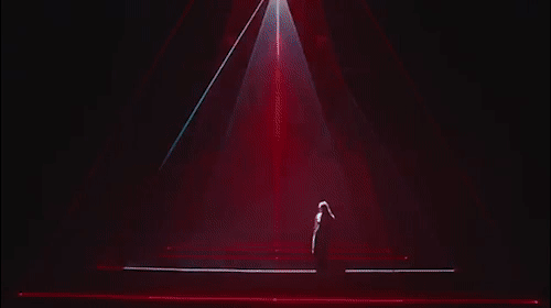 Solo singer silhouetted on a stage with dynamic red and white stage lighting creating a powerful spotlight effect in the dark.