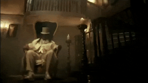 Man in period clothing sitting in an armchair in a dimly lit room with candle and staircase, giving a vintage, eerie feel.