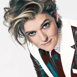 Close-up portrait of a woman with short blonde-streaked hair wearing a white jacket and pinstripe shirt against a seamless white background.