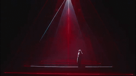 Figure standing inside a large triangular pyramid structure illuminated by stark red laser beams and ambient stage fog.