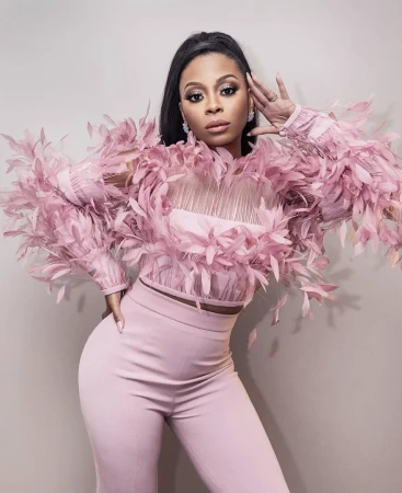 Attractive woman poses in a pink feathered top and matching slacks against a neutral background in a full-body shot with soft lighting.