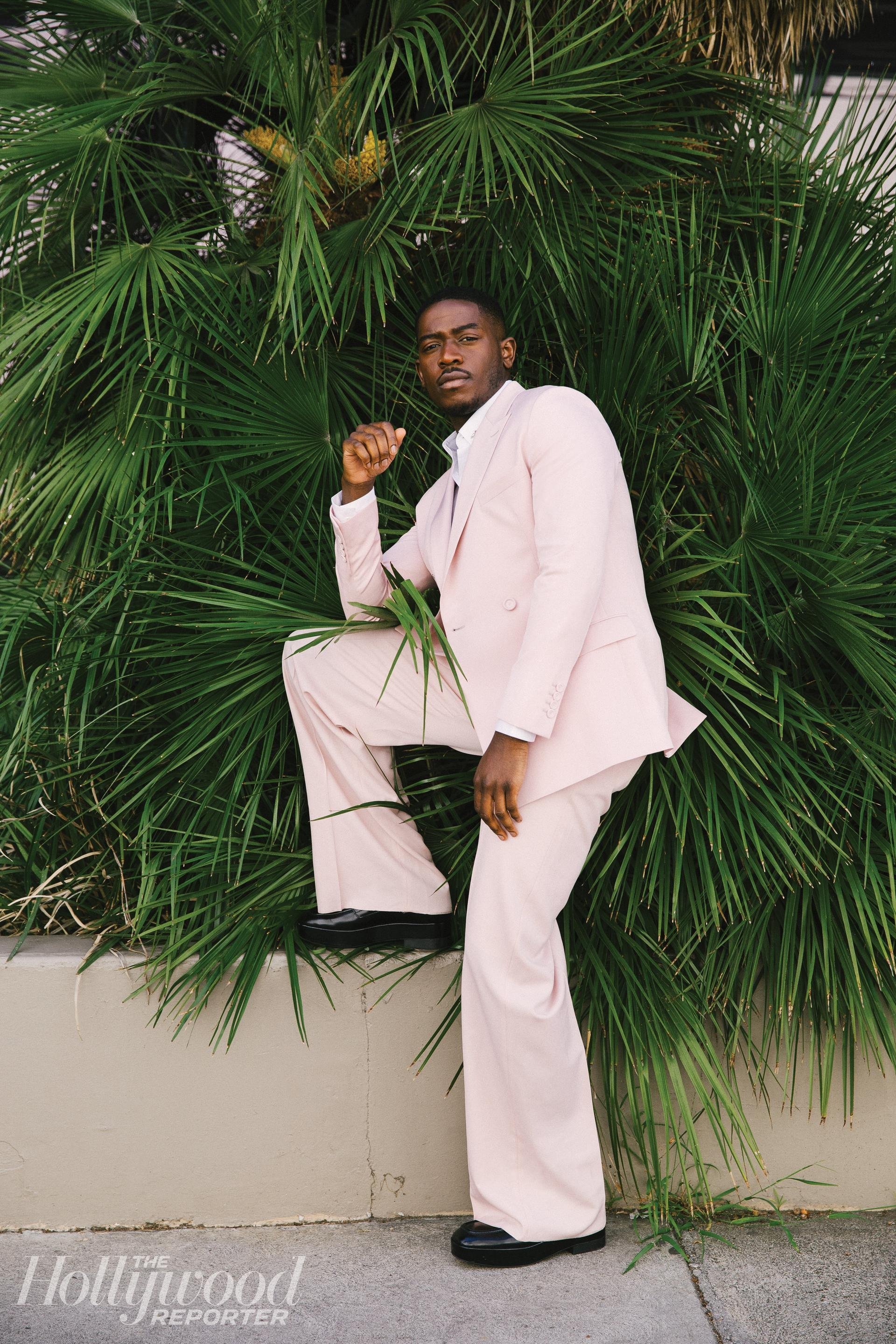 Portrait of a Black male in a pink suit posing against a leafy green plant wall, exuding a stylish and confident presence.