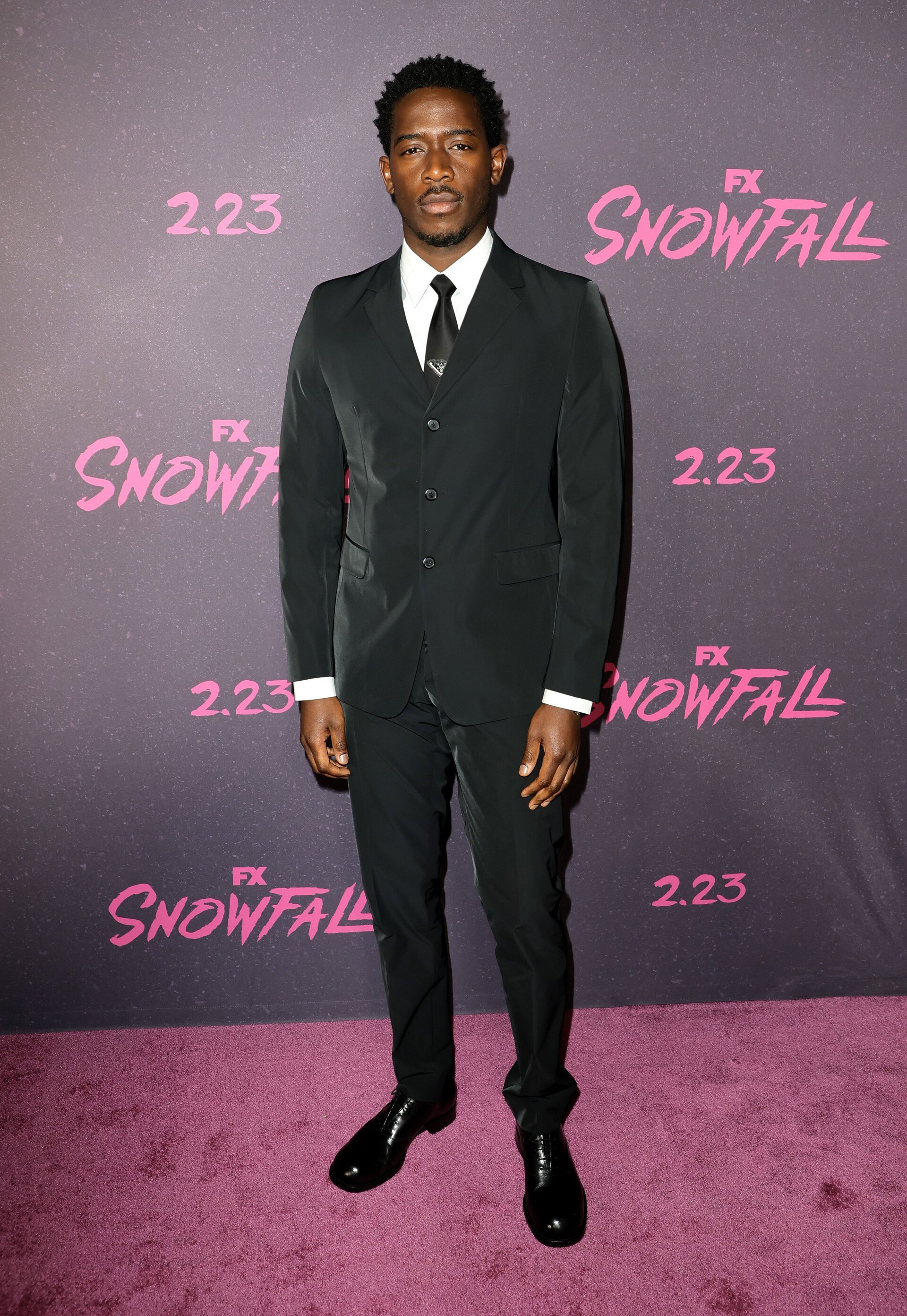 Full-length portrait of a Black man wearing a suit and tie standing on a purple carpet at a red carpet event with 'Snowfall' text in background.