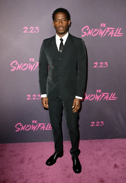 Full-length portrait of a Black man wearing a suit and tie standing on a purple carpet at a red carpet event with 'Snowfall' text in background.