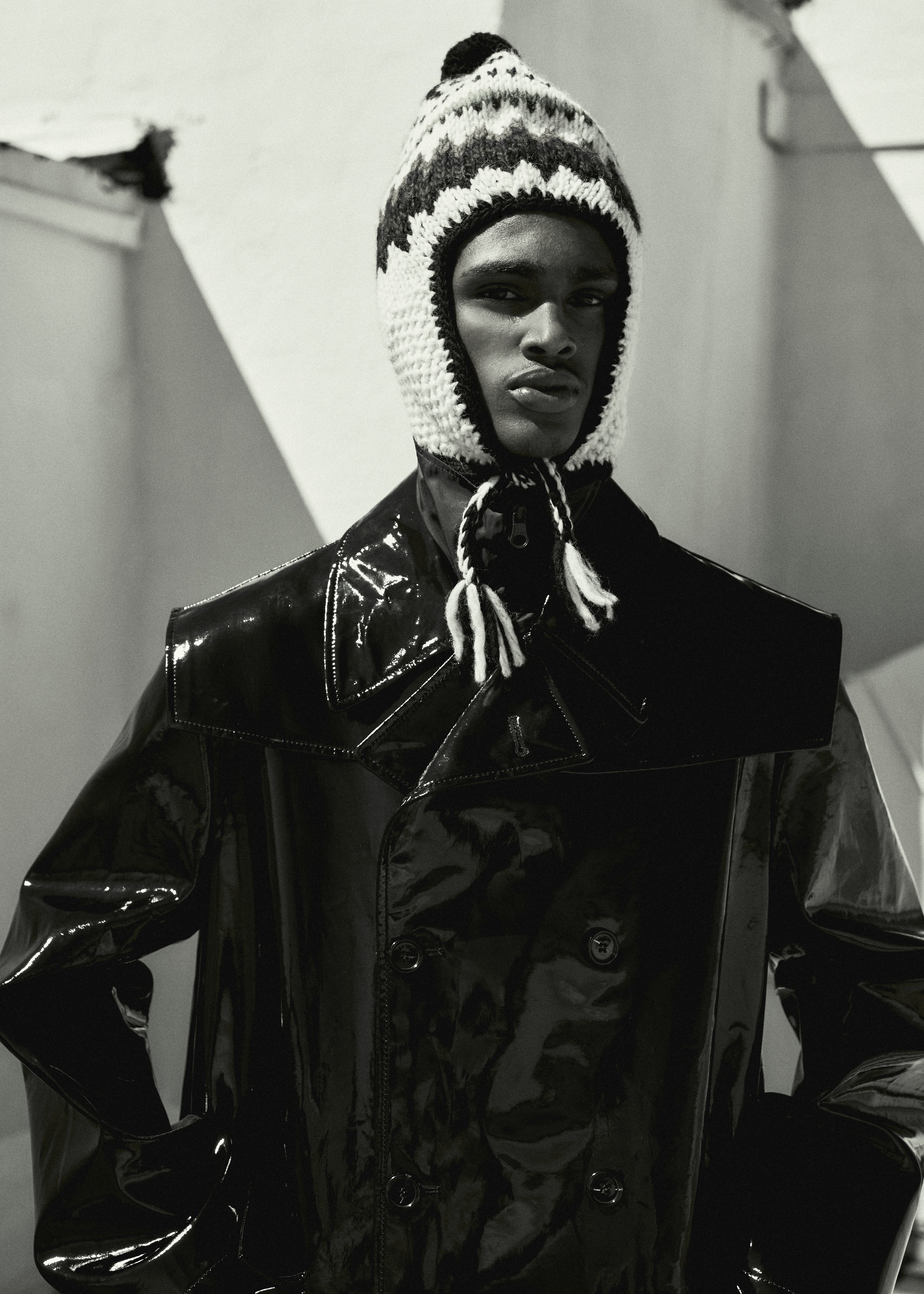Monochromatic portrait of a dark-skinned adult male in a knit cap with tassels, shiny black jacket, against an abstract background.