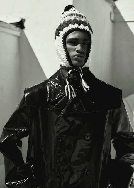 Monochromatic portrait of a dark-skinned adult male in a knit cap with tassels, shiny black jacket, against an abstract background.