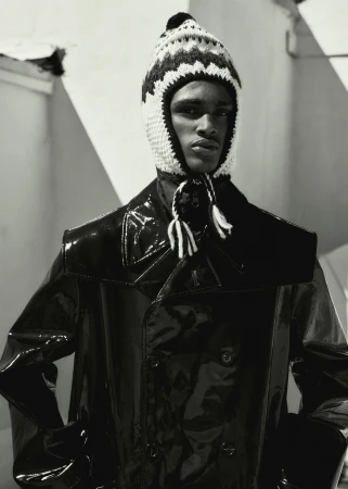 Monochromatic portrait of a dark-skinned adult male in a knit cap with tassels, shiny black jacket, against an abstract background.