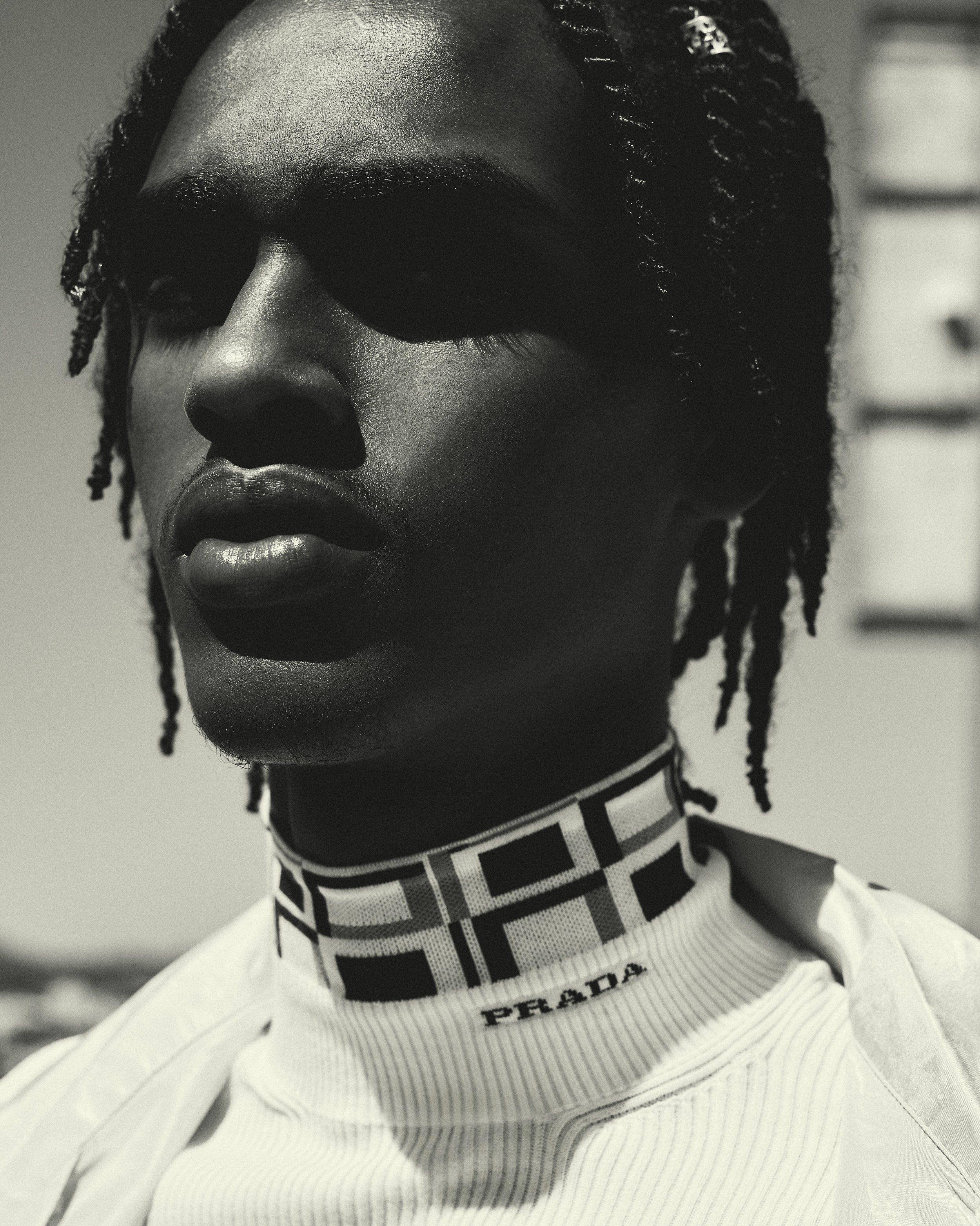 Close-up portrait of a Black male model with braided hair wearing a Prada turtleneck in an outdoor setting, showcasing fashion and style.