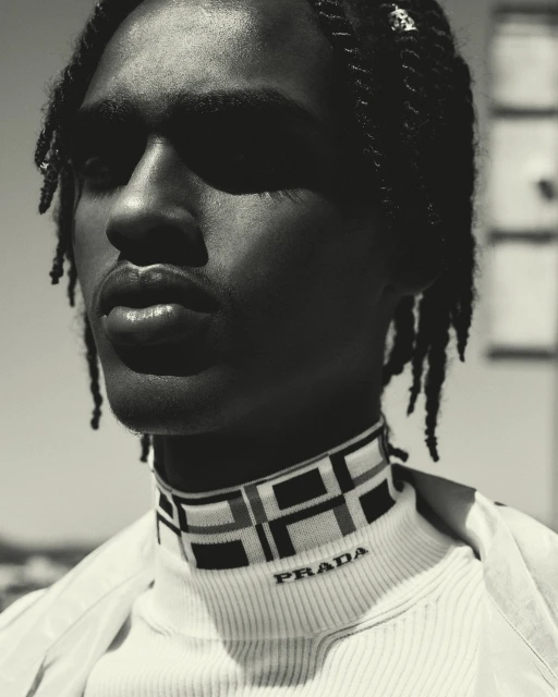 Close-up portrait of a Black male model with braided hair wearing a Prada turtleneck in an outdoor setting, showcasing fashion and style.