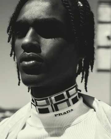 Close-up portrait of a Black male model with braided hair wearing a Prada turtleneck in an outdoor setting, showcasing fashion and style.