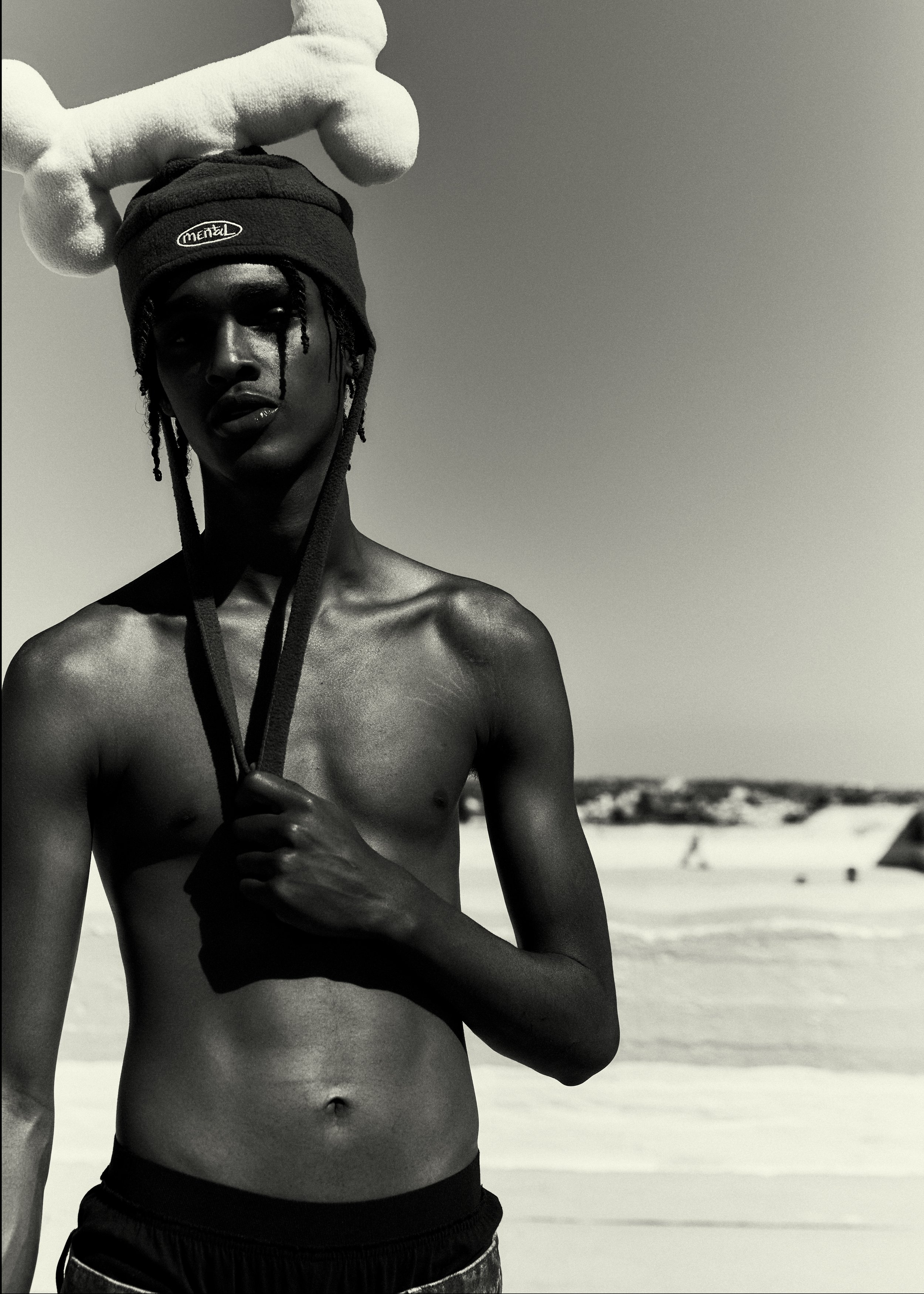 Monochrome portrait of a shirtless adult male on a beach, wearing a beanie with a plush toy bone, against a sunny sky.