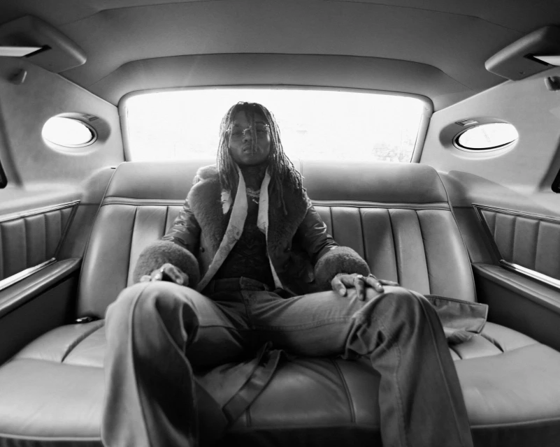 Black and white photograph of a man with locs sitting in the back of a limousine, wearing a coat, looking directly at the camera.