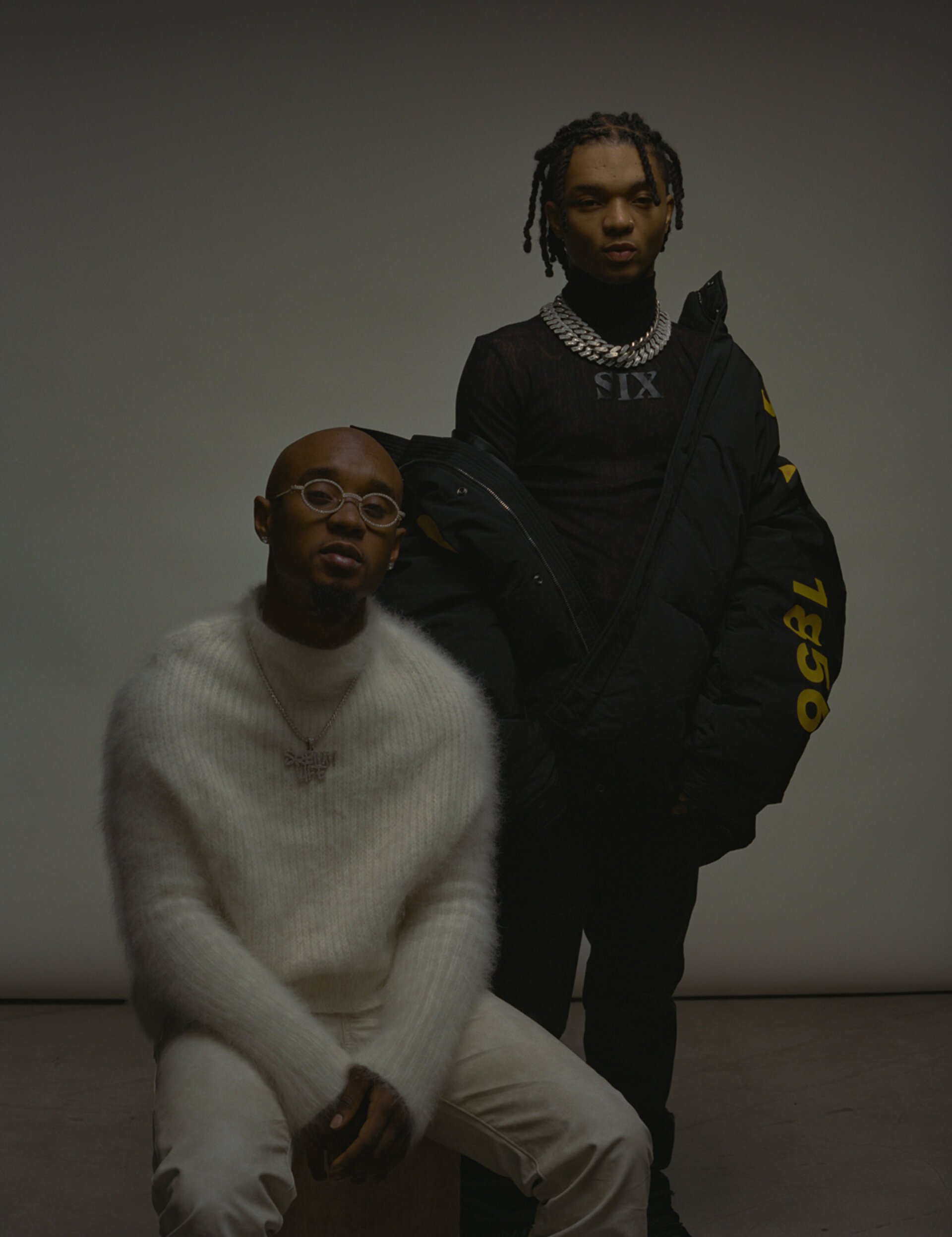 Two adult men pose indoors against a grey background. One is seated, wearing glasses and a white sweater; the other is standing, wearing black.