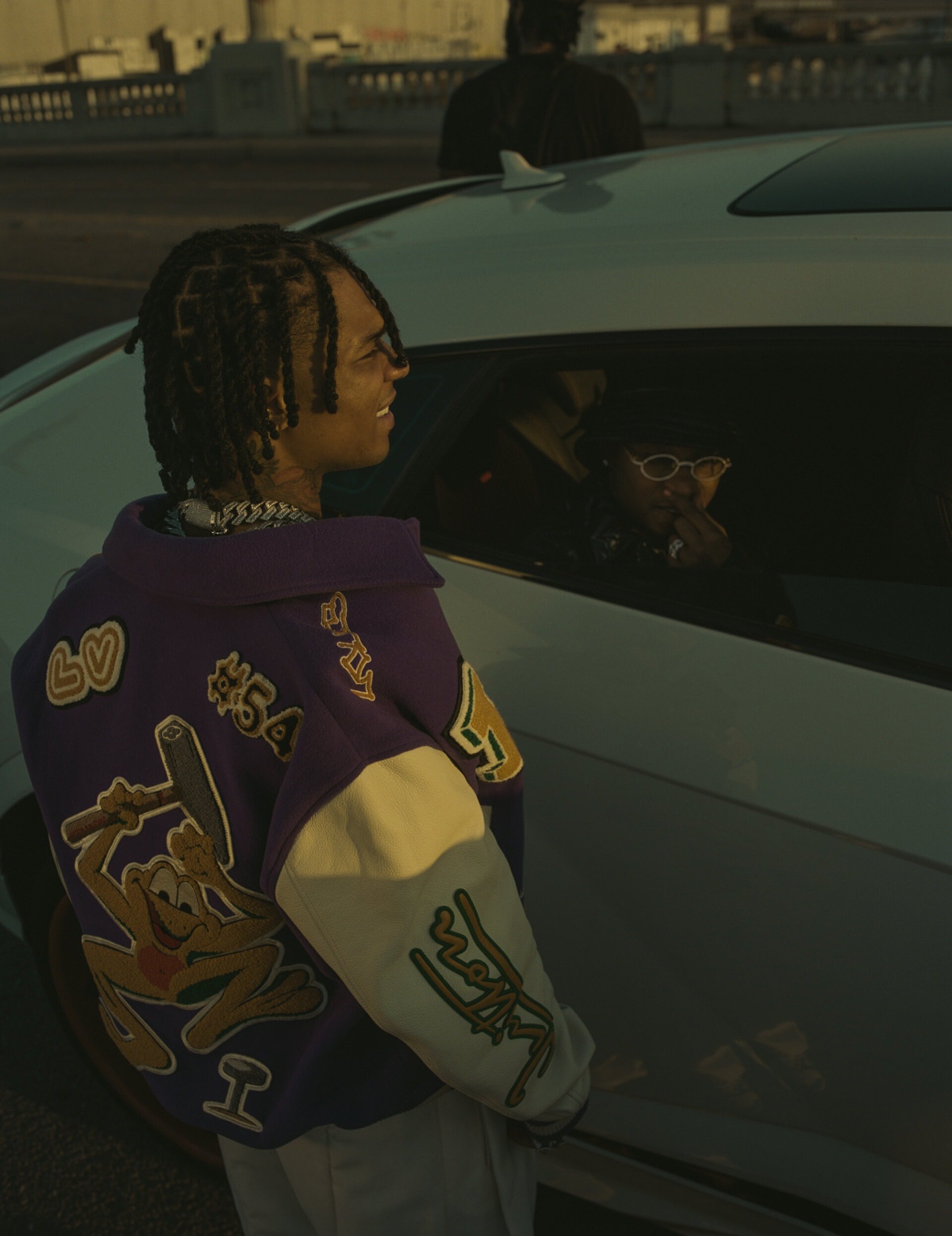 Man in Los Angeles Lakers jacket standing near a car at dusk. He is smiling and wears his hair in dreadlocks with a chain.