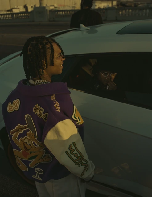 Man in Los Angeles Lakers jacket standing near a car at dusk. He is smiling and wears his hair in dreadlocks with a chain.