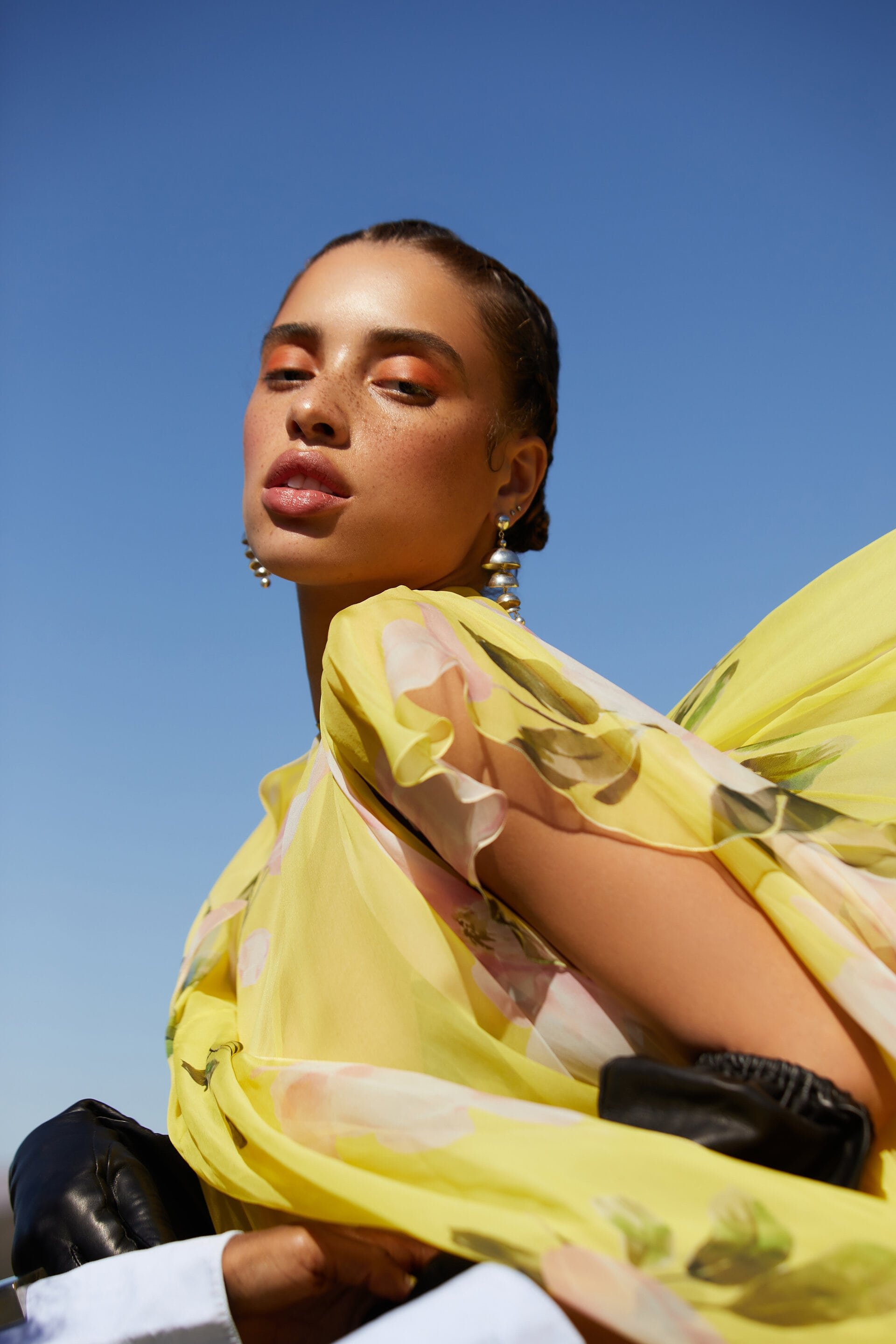 Woman in yellow floral dress and pearl earrings poses against a clear blue sky in a fashion editorial portrait with low camera angle.
