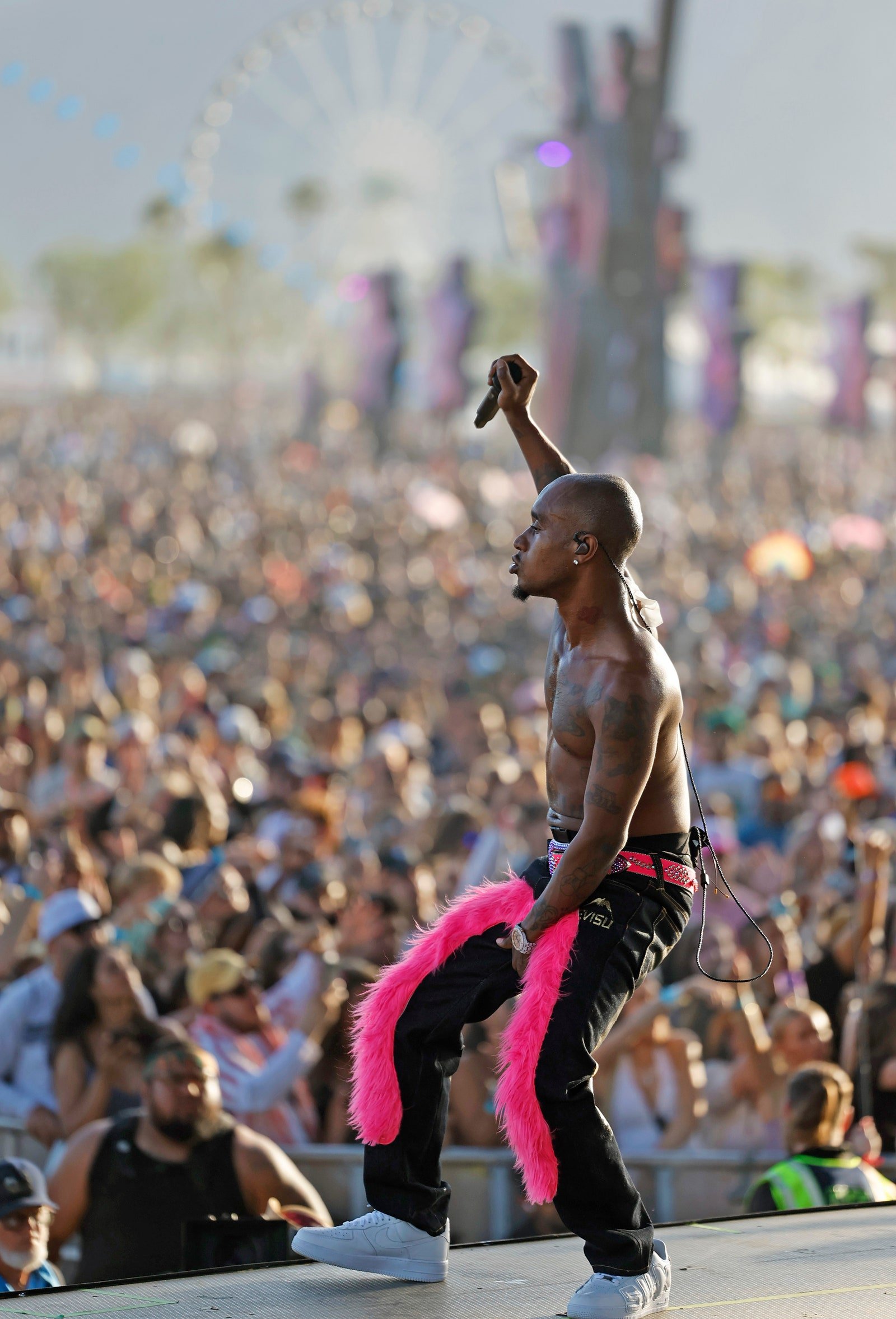 African American male musician performs on stage at outdoor music festival in black pants with pink feather trim and white sneakers.