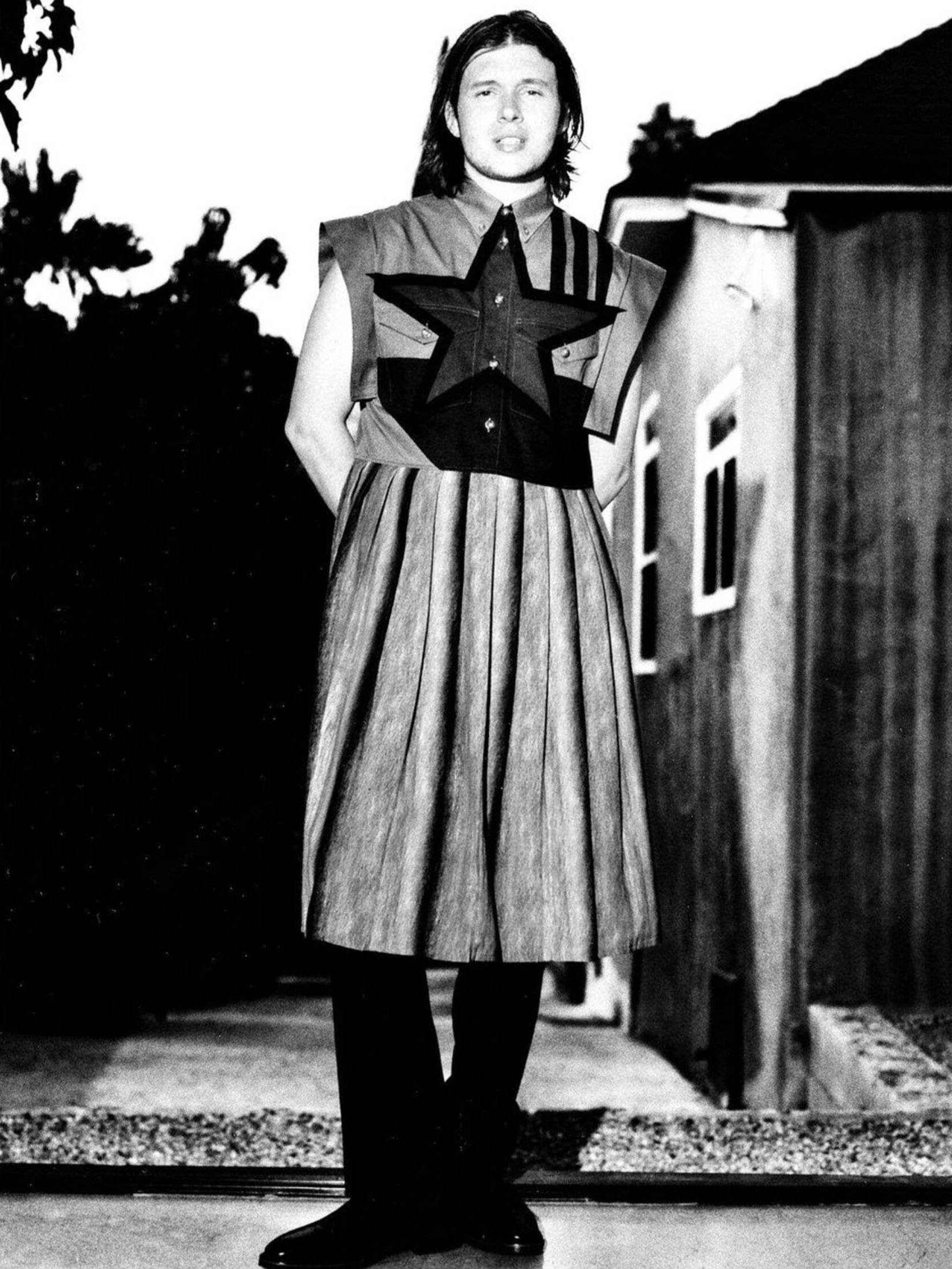 Person in fashion attire stands in front of house and tree, outdoors in black and white. Shirt with star accent and pleated skirt.