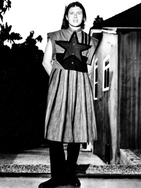 Person in fashion attire stands in front of house and tree, outdoors in black and white. Shirt with star accent and pleated skirt.