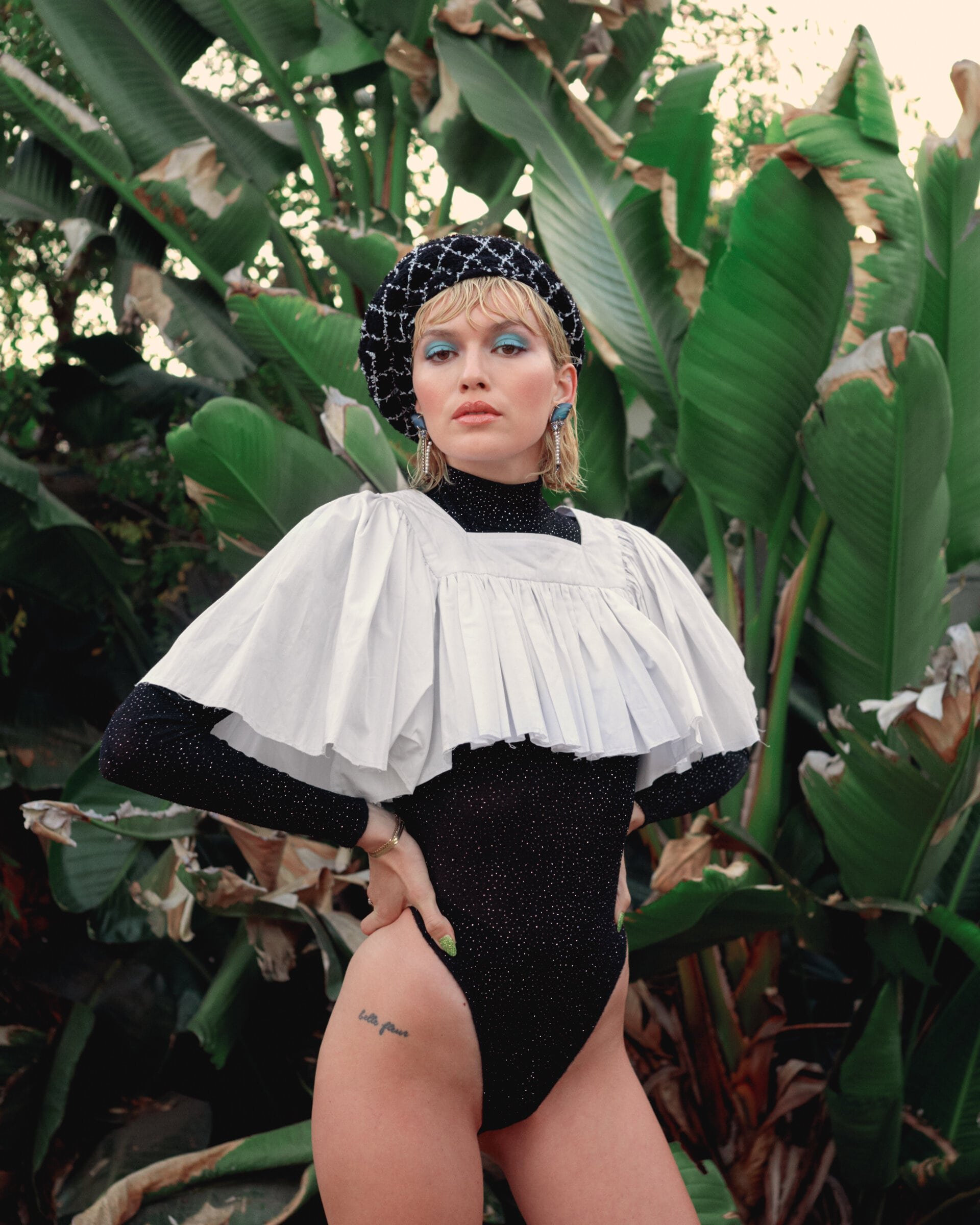 A woman poses outdoors in a black and white bodysuit with puffed sleeves, wearing a beret, with large green tropical plants in the background.