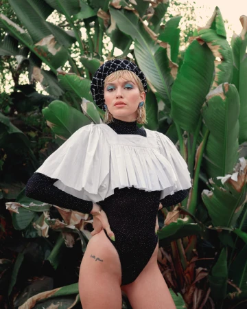 Model wearing a black sparkly bodysuit, pleated white overlay collar, and checkered beret in front of lush tropical foliage