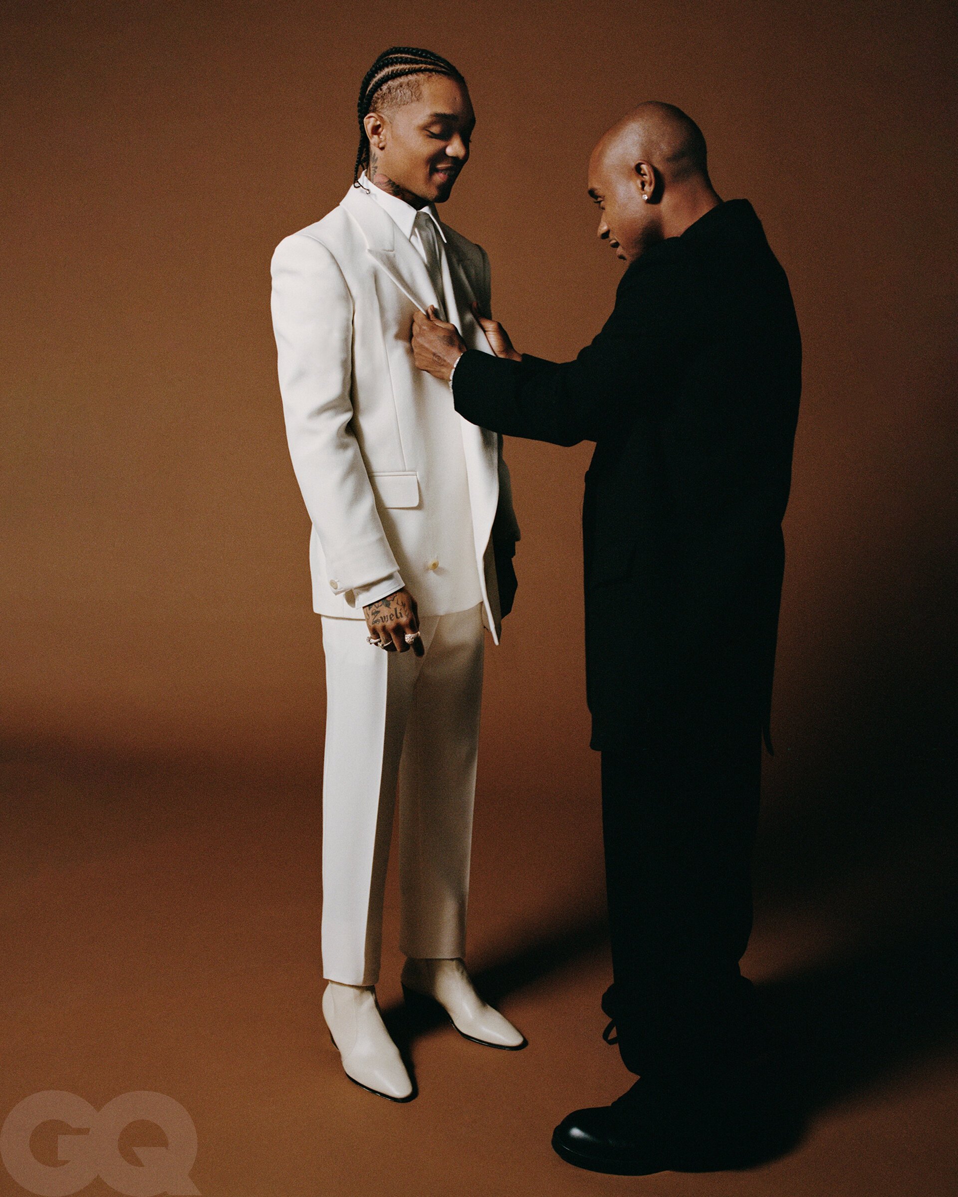 Two men in suits. One man in a white suit is being adjusted by another man in a black suit, set against a warm brown backdrop.
