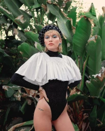 Model wearing a black sparkly bodysuit, pleated white overlay collar, and checkered beret in front of lush tropical foliage