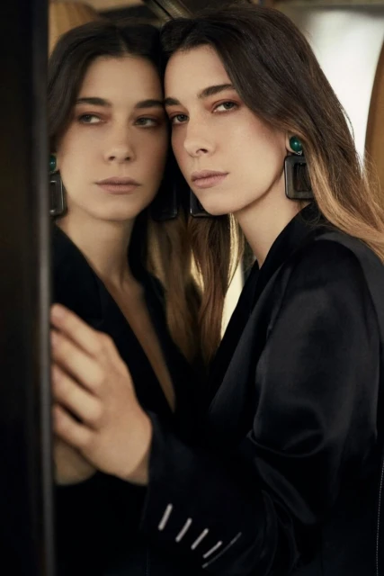 Young woman in a black jacket gazes seriously at the camera with a mirrored reflection, highlighting her square earrings.