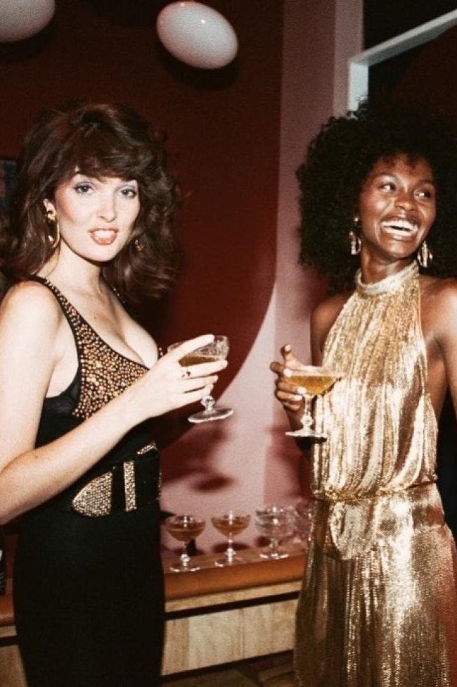 Two women in elegant dresses toast with champagne glasses at an indoor event, lit by an overhead lamp and soft lighting.