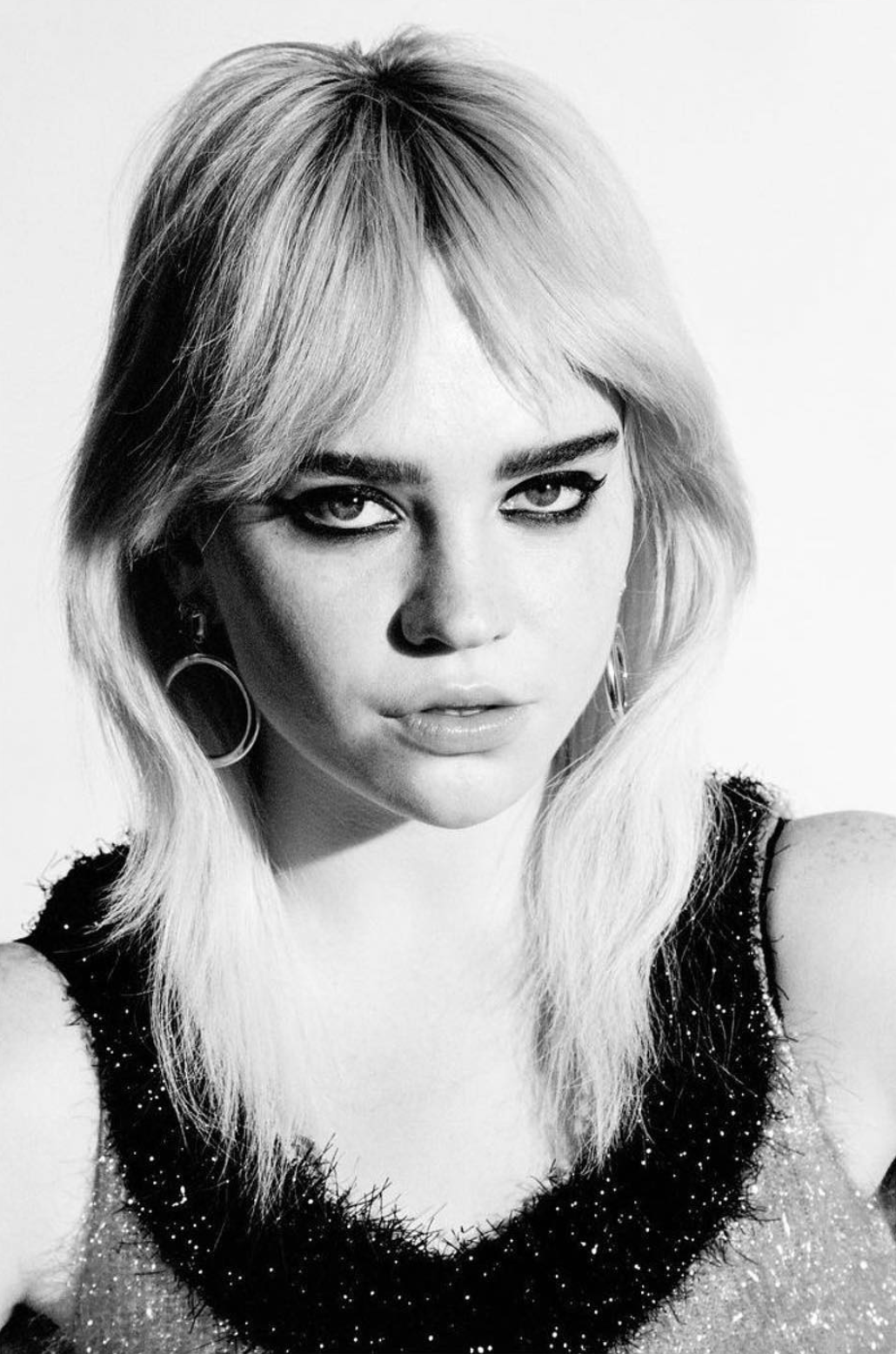 Black and white portrait of an adult woman with blond hair, dark eye makeup, and hoop earrings against a plain white background.