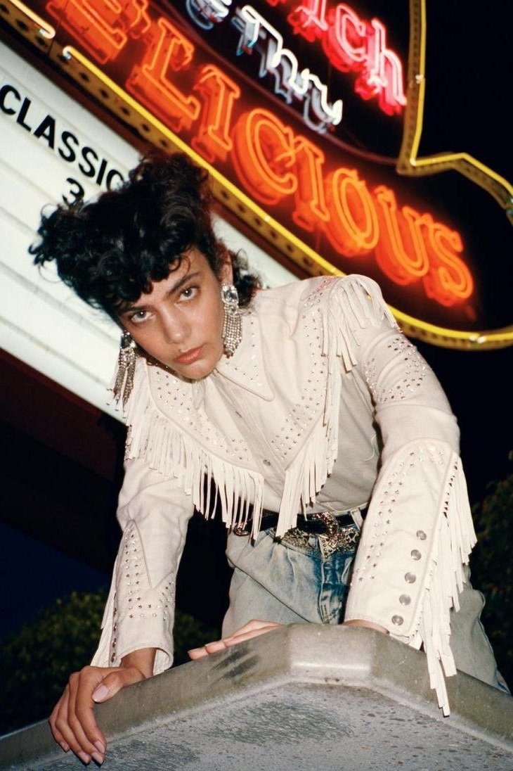 Woman in fringe jacket poses in front of neon 'Delicious' sign at night. She wears jeans and ornate earrings, styled with an upward gaze.