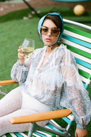 A woman with sunglasses and a headscarf sits in a beach chair, holding a wine glass on the lawn, looking directly at the camera.