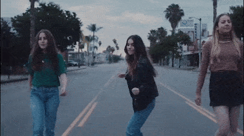 Three adult women walk across a street in an urban area with palm trees and buildings in the background under an overcast sky.